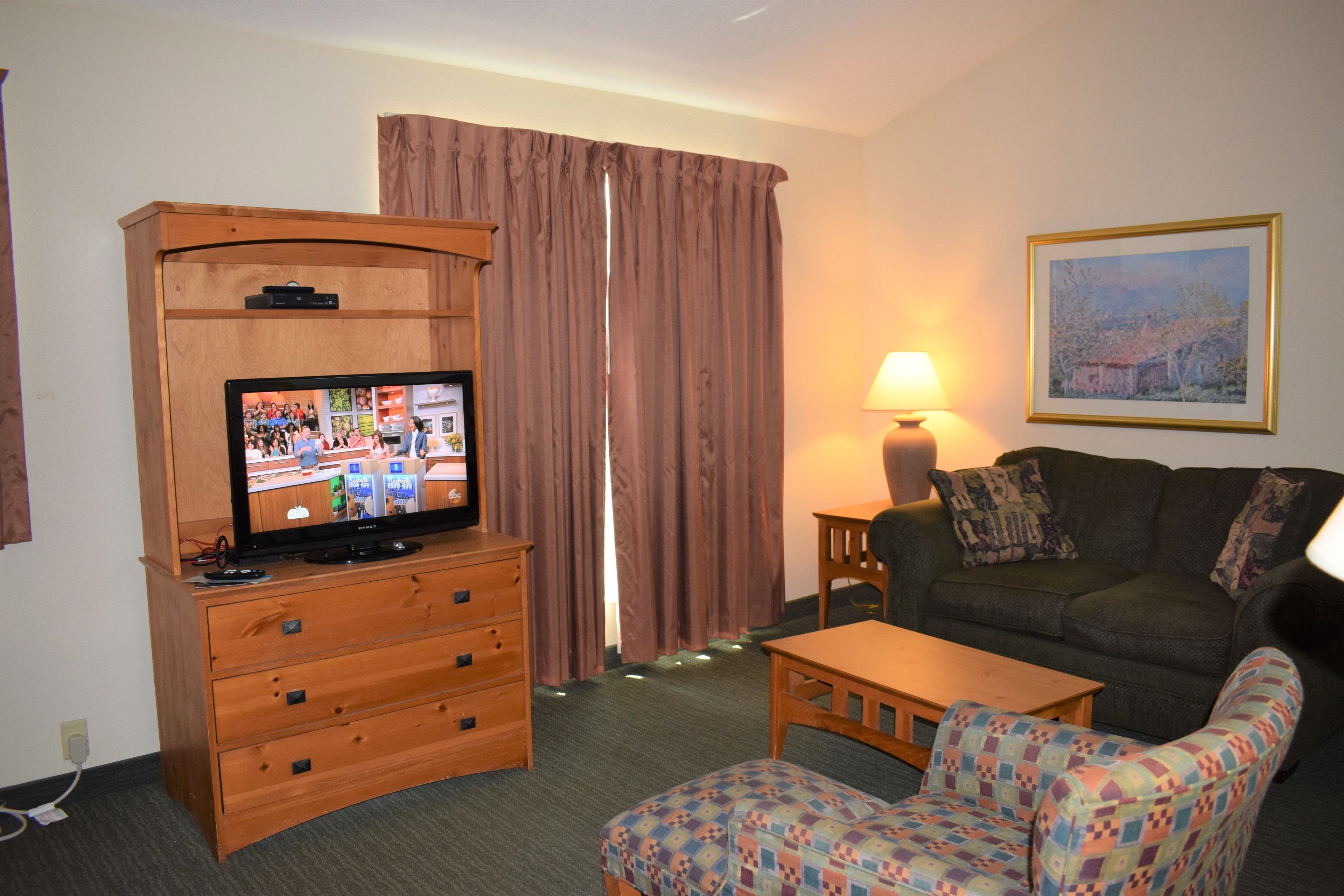 A room with a television on a wooden cabinet, a sofa, a patterned armchair, a table, and a lamp.