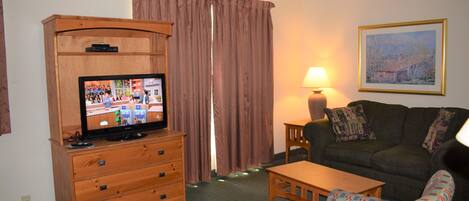 Ein Zimmer mit Fernseher auf einer Holzkommode, einer Couch, einem gemusterten Sessel, einem Tisch und einer Lampe.