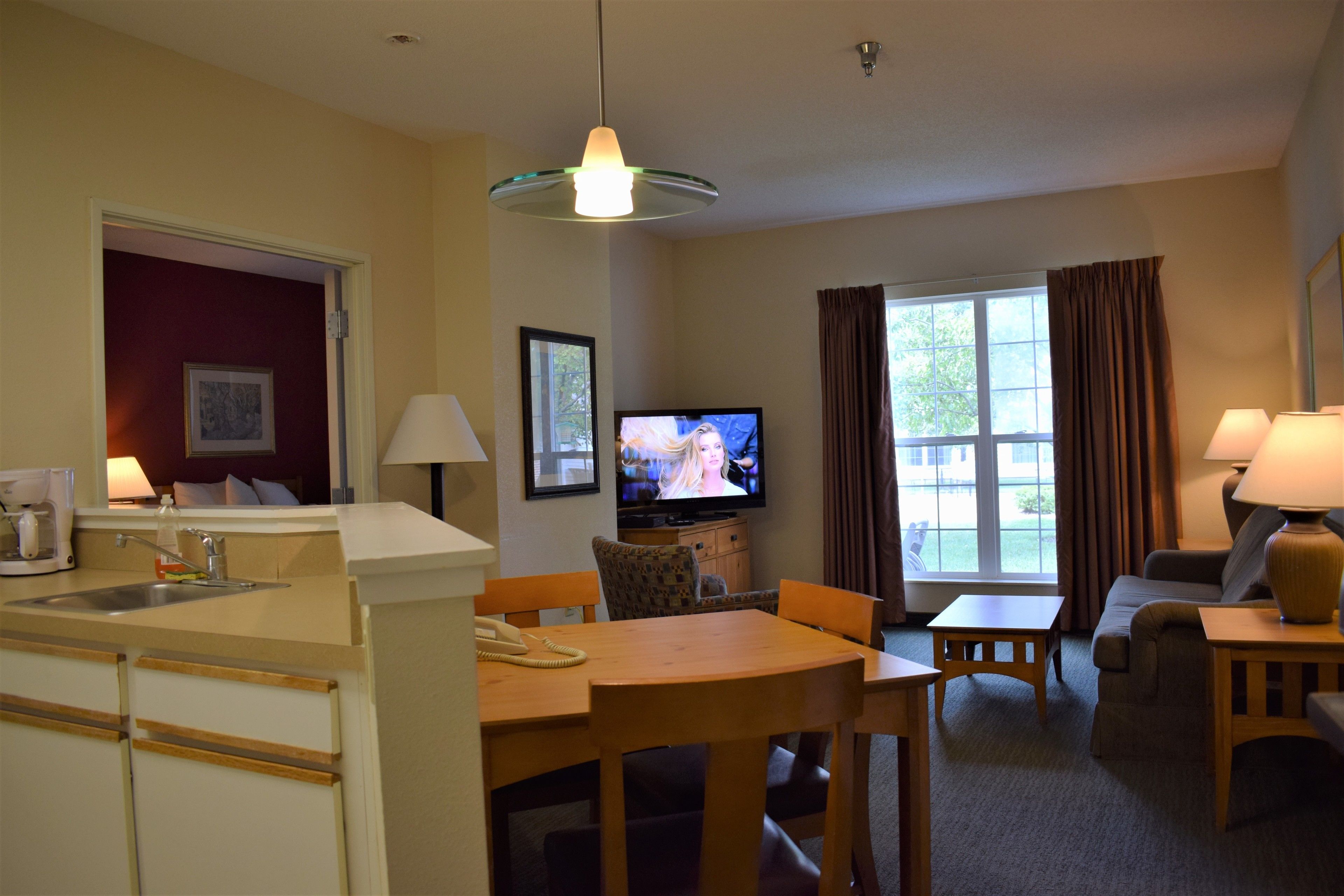 A hotel room with a kitchenette, a dining area, a living room with a sofa and TV, and a large window with curtains.