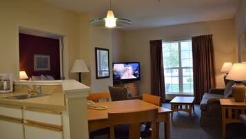 A hotel room with a kitchenette, a dining area, a living room with a sofa and TV, and a large window with curtains.