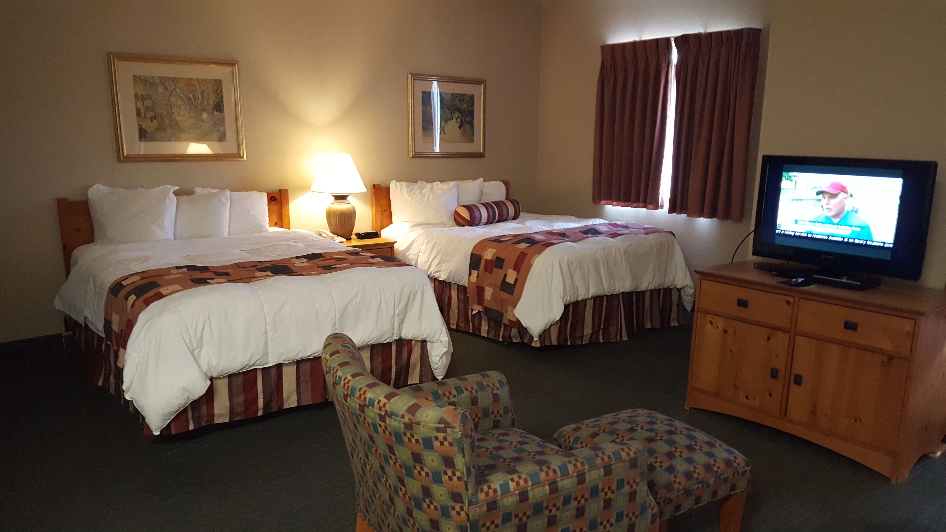 A hotel room with two beds, a television, a wooden cabinet, and a patterned armchair.