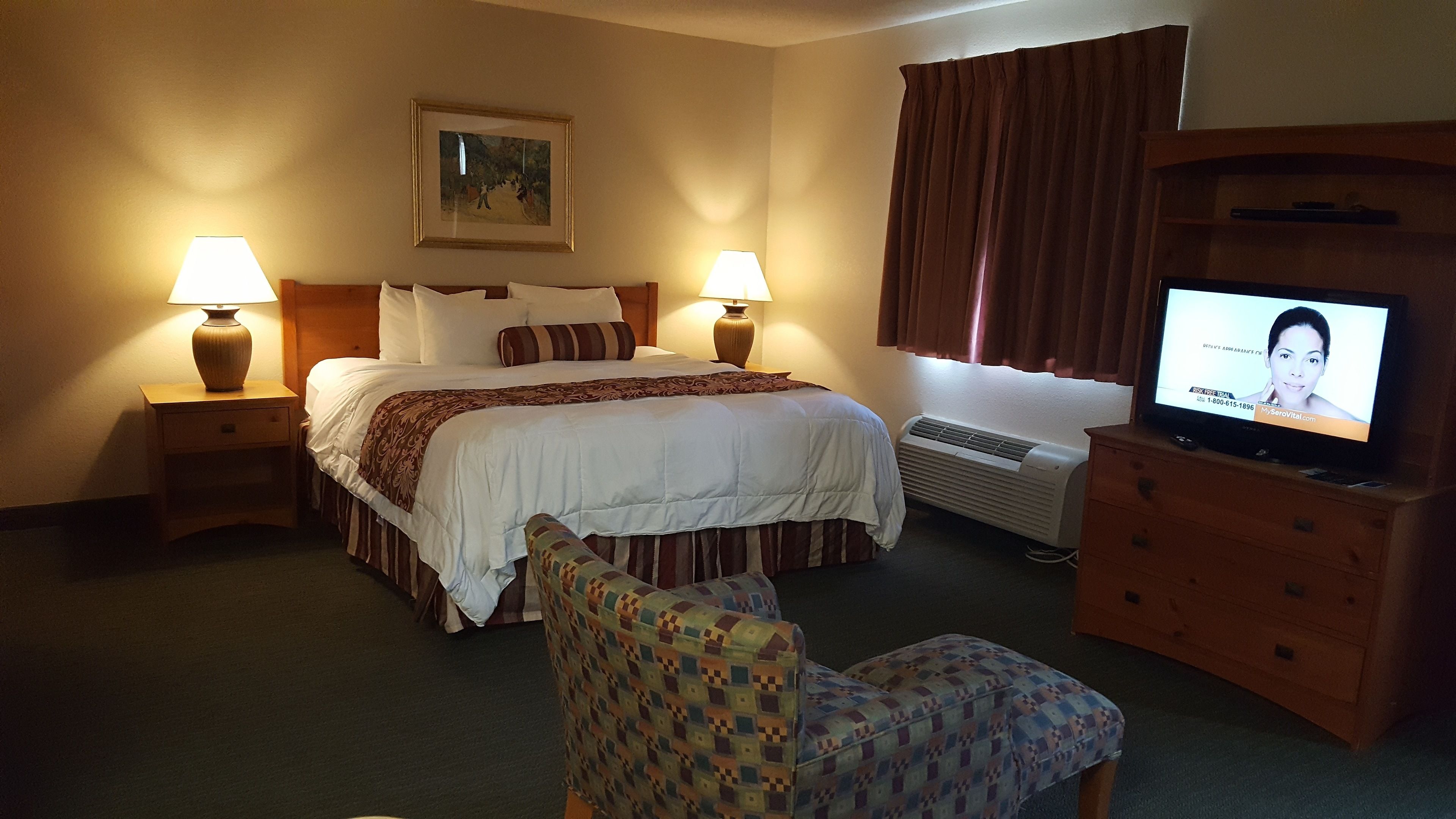 A hotel room with a bed, two bedside lamps, a television, a patterned armchair, a dresser, and a painting on the wall.