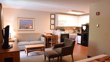 A compact living space with a sofa, armchair, dining table, kitchenette, and a painting on the wall.