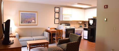 A compact living space with a sofa, armchair, dining table, kitchenette, and a painting on the wall.