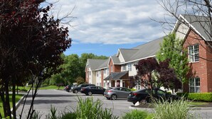 A parking lot with several cars, a brick building with multiple windows, and surrounding greenery. - Cresthill Suites Syracuse (East Syracuse)