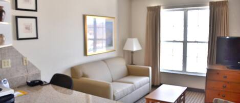 A hotel room with a kitchenette, a sofa, a dining table, a TV, and a window with curtains.