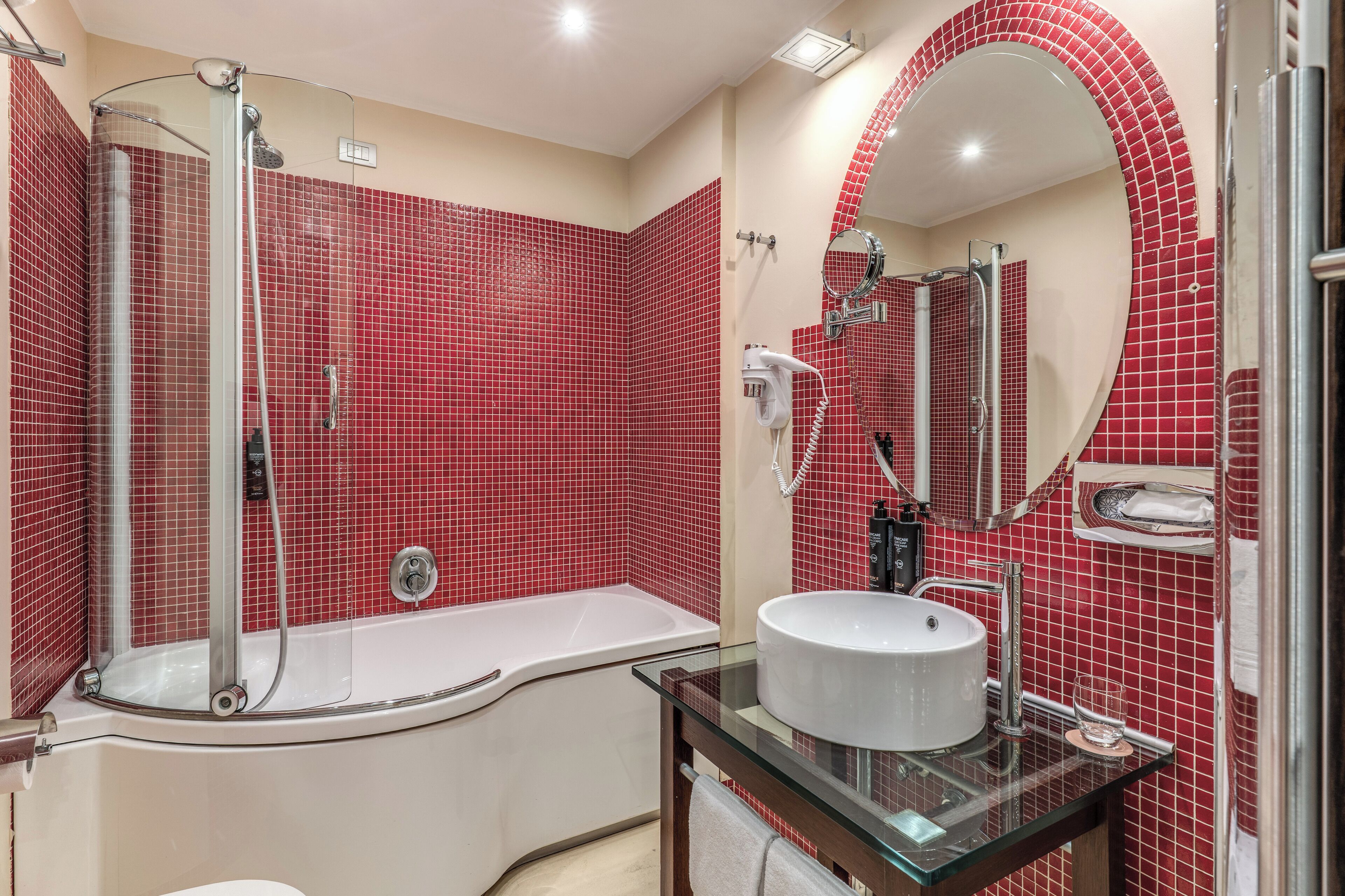 A bathroom with a glass-enclosed shower, a white bathtub, and a round sink with a glass countertop.