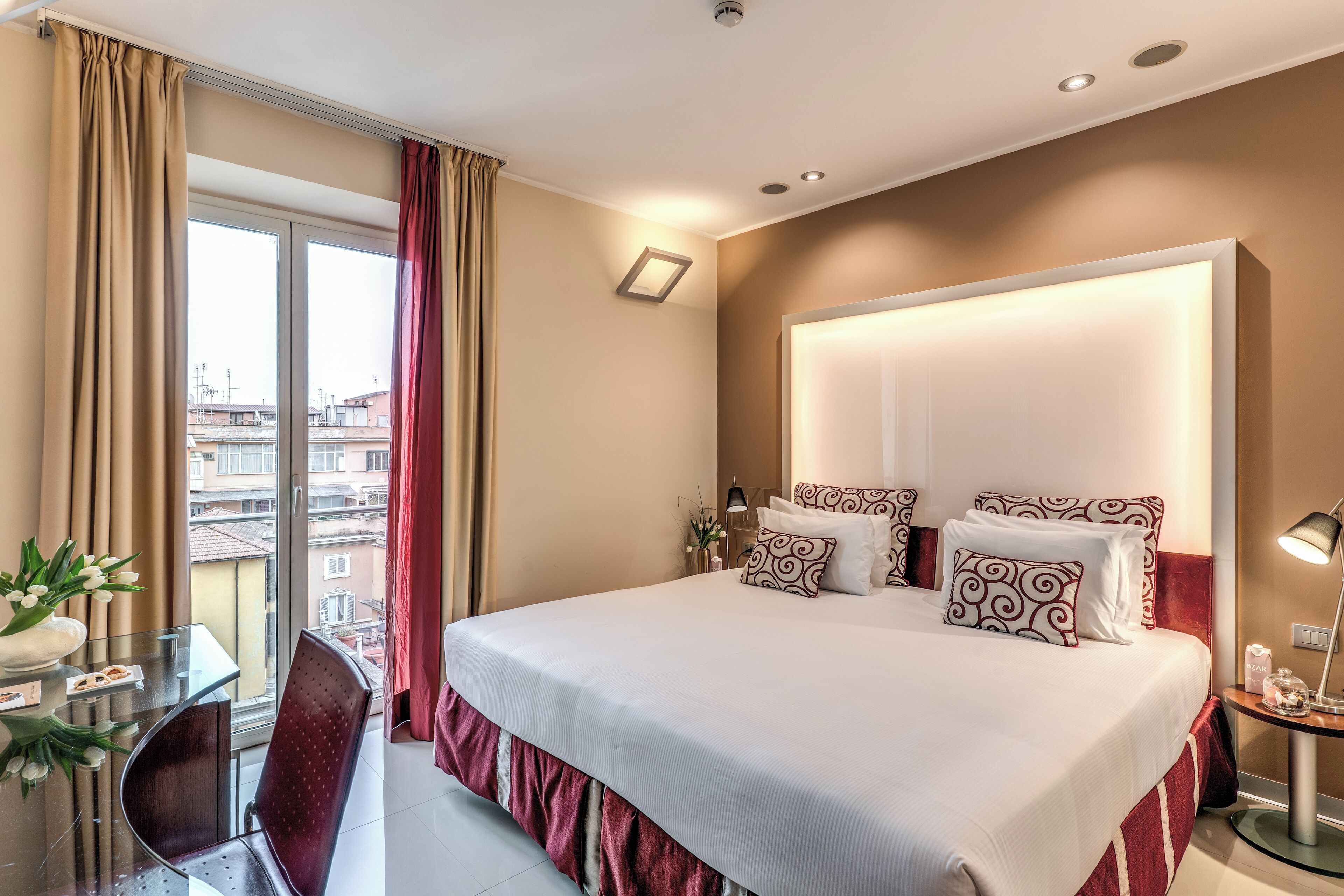 A hotel room with a large bed, a desk with a chair, a window with curtains, and a bedside table with a lamp.