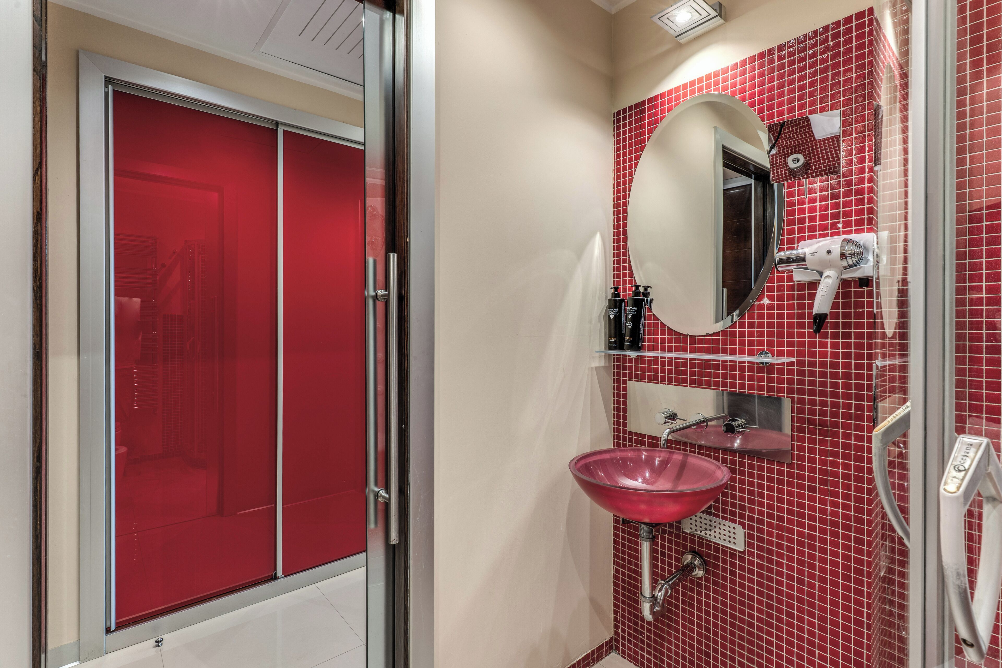 A bathroom with a red tiled wall, a white sink, a mirror, and a hairdryer mounted on the wall.