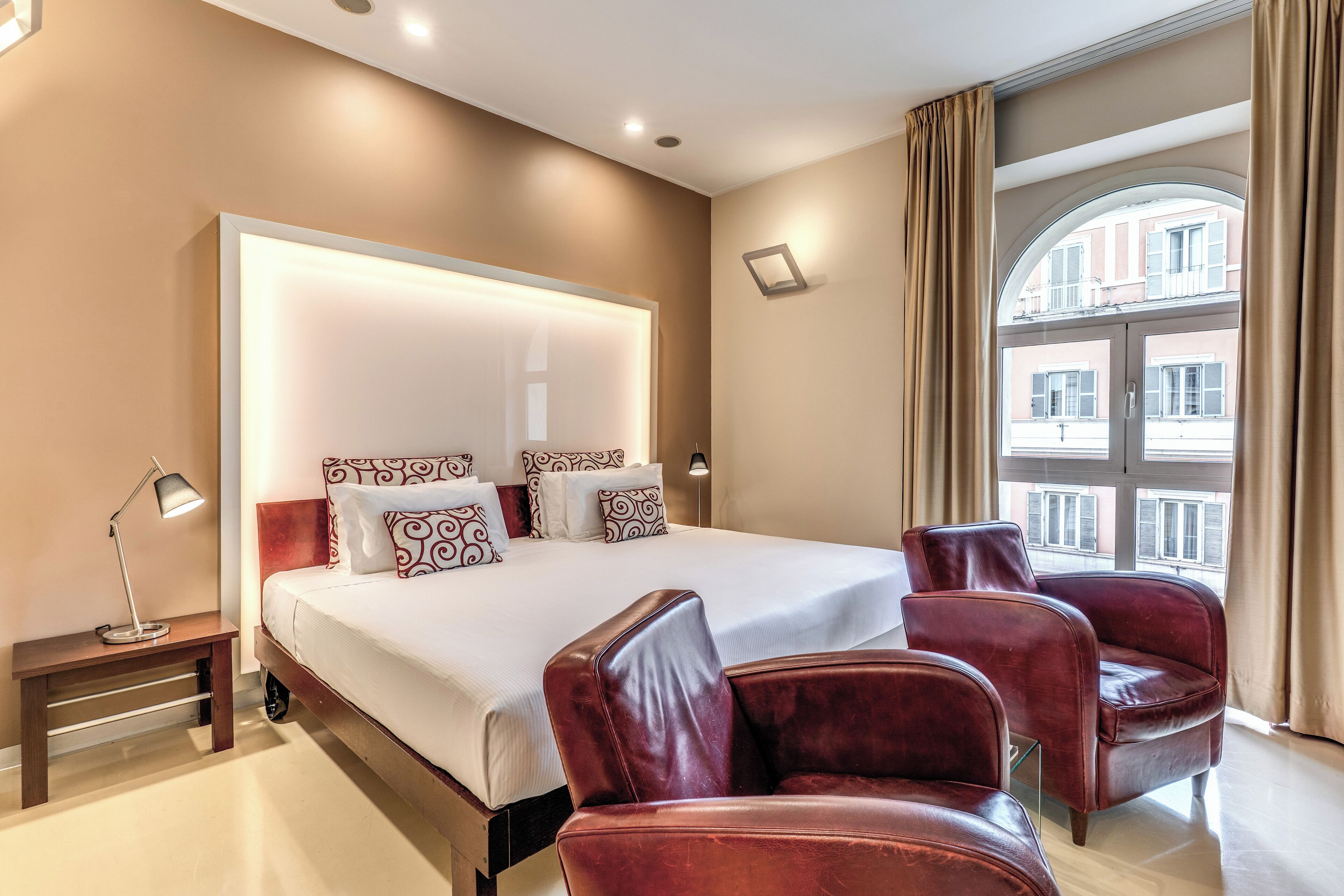 Une chambre d’hôtel avec un grand lit, deux fauteuils en cuir, une table de chevet avec une lampe et une vue sur les bâtiments par la fenêtre.