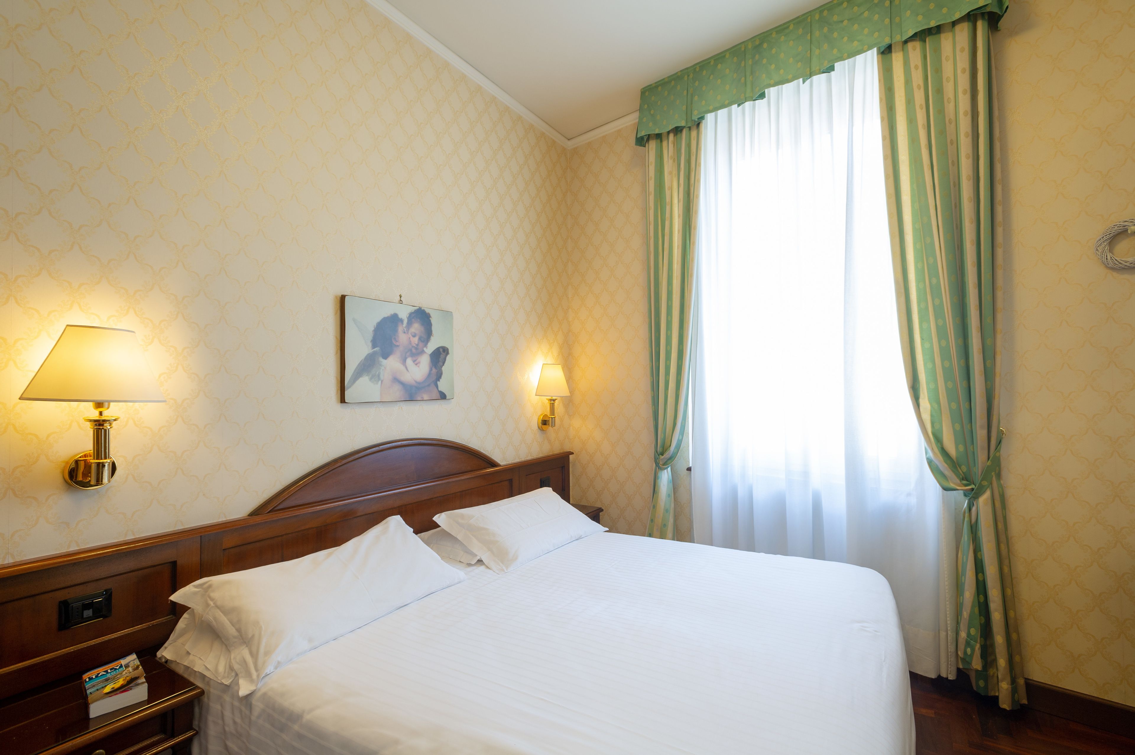 A neatly made bed with white linens, two bedside lamps, a framed picture on the wall, and green-striped curtains on a window.