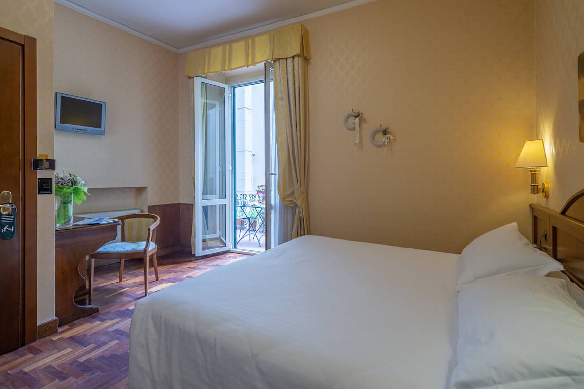 A hotel room with a bed, a desk with a chair, a television, and a balcony with curtains.