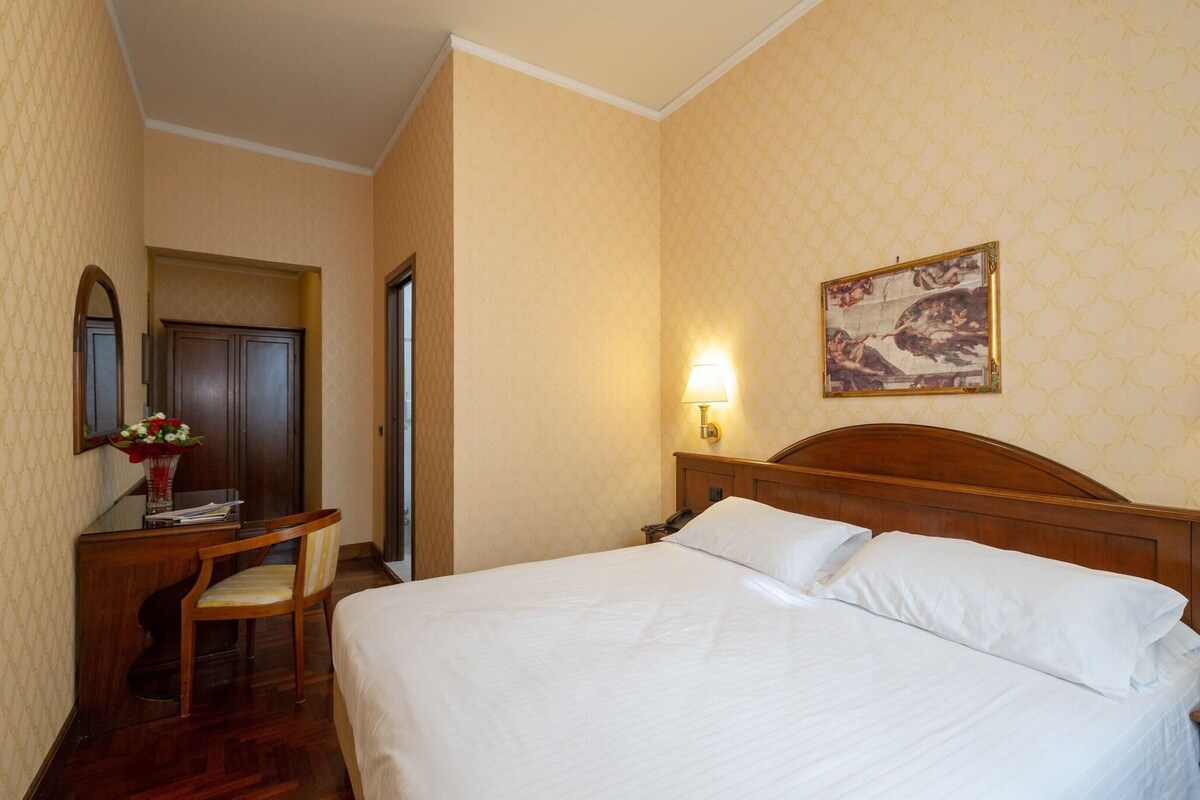 A hotel room with a bed, a desk with a chair, a mirror, and a wall-mounted lamp.