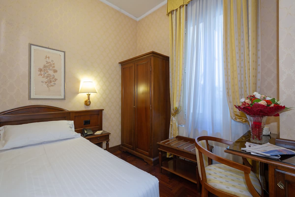 A hotel room with a bed, a wooden wardrobe, a desk with a chair, a lamp, and a vase with flowers.