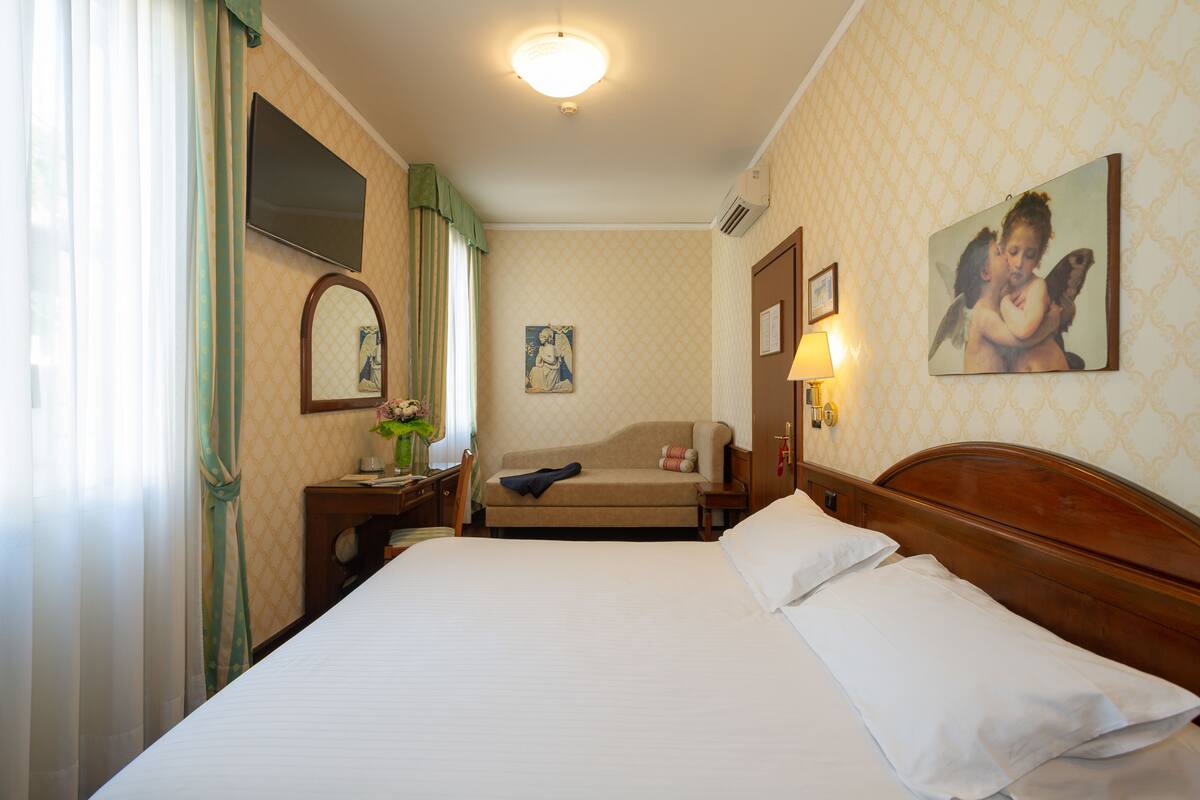 A hotel room with a large bed, a sofa, a desk with a lamp, a painting on the wall, and a window with curtains.