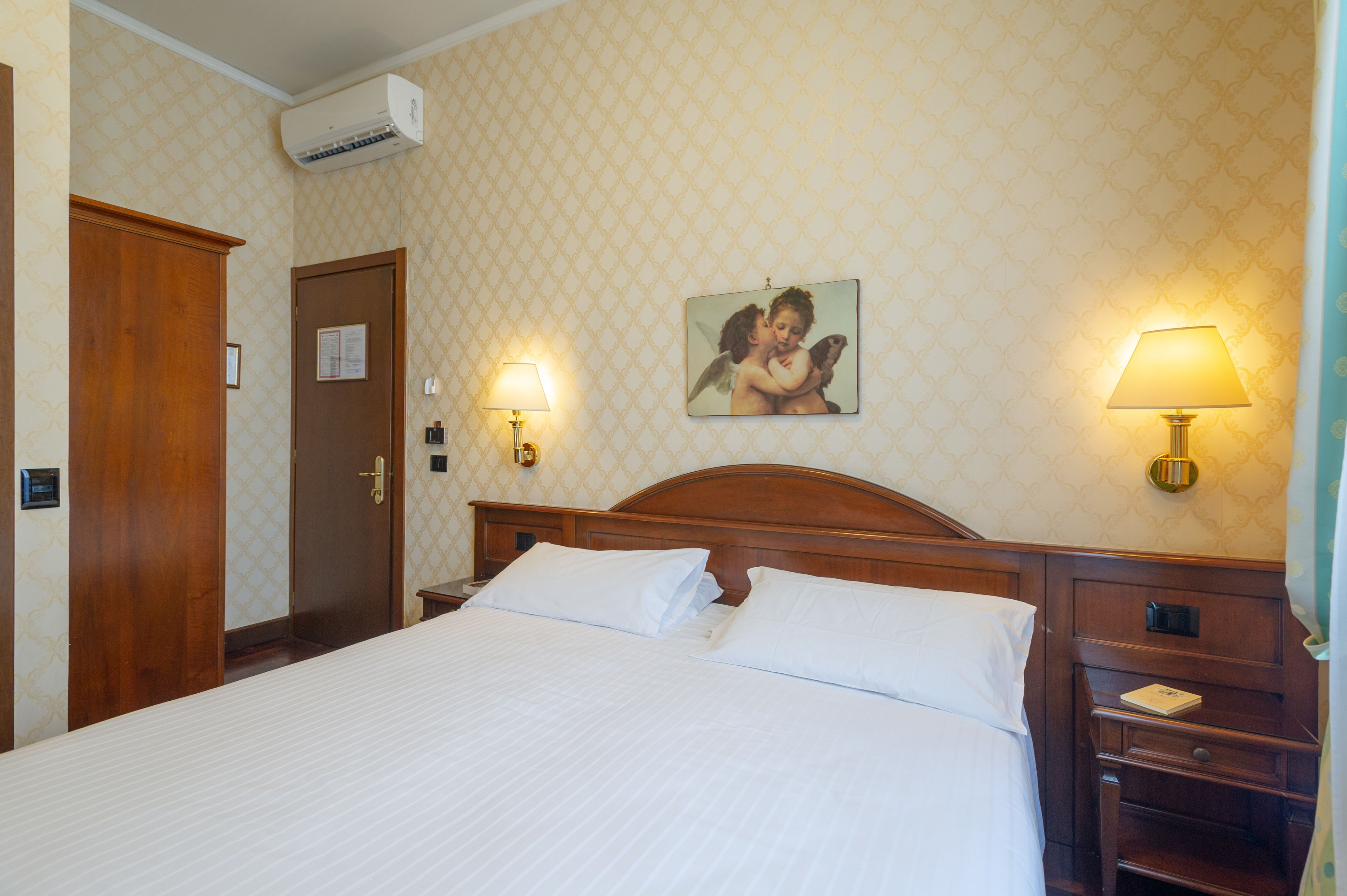 A hotel room with a large bed, wooden headboard, two bedside lamps, a nightstand, and a wall-mounted air conditioner.