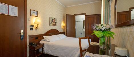 A hotel room with a bed, bedside table, lamp, mirror, and a vase with flowers.