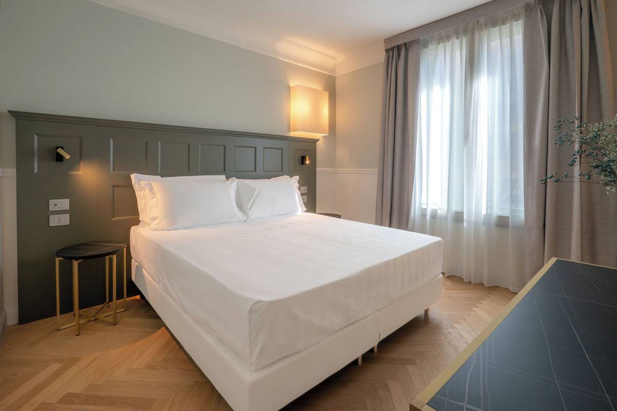Ein Hotelzimmer mit einem großen Bett, einem kleinen Beistelltisch, einem Nachttisch, einer Lampe und einem Fenster mit Vorhängen.