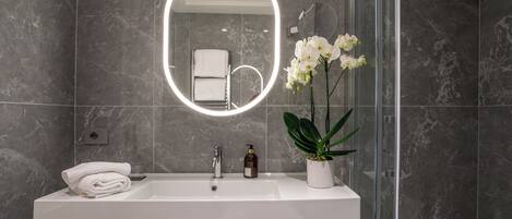 A modern bathroom with a white sink, a mirror with a circular light, and a potted orchid.