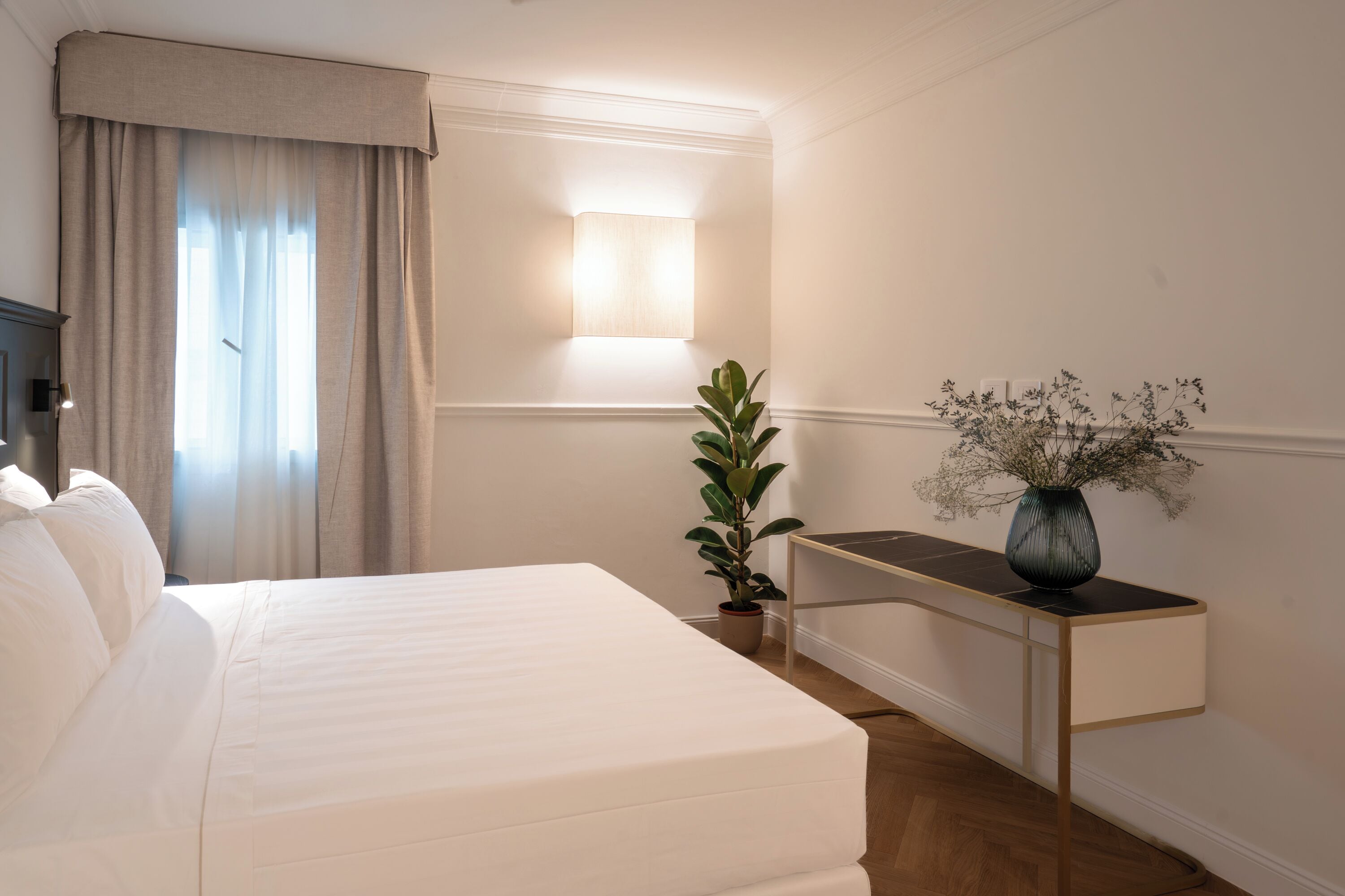 Habitación de hotel con una cama grande, una mesita de noche con un florero y una planta en maceta, además de una lámpara fijada a la pared.