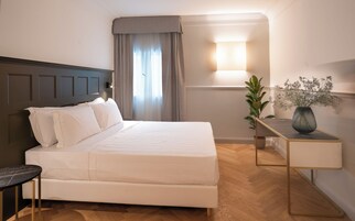 Un letto rifatto con lenzuola bianche, un comodino con un vaso di fiori e una lampada a parete.