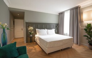 Una habitación de hotel con una cama grande, una mesita de noche con un florero, un sillón verde y una planta en maceta.