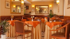 A restaurant with multiple tables covered with orange tablecloths, wooden chairs, and potted plants.