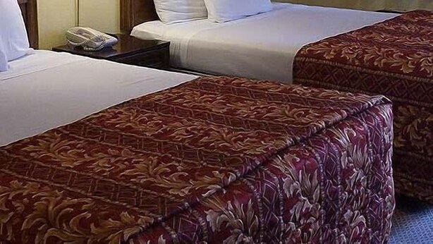 A hotel room with two beds, each with white linens and a brown headboard. There are two bedside lamps, a framed picture on the wall, and a window with curtains.