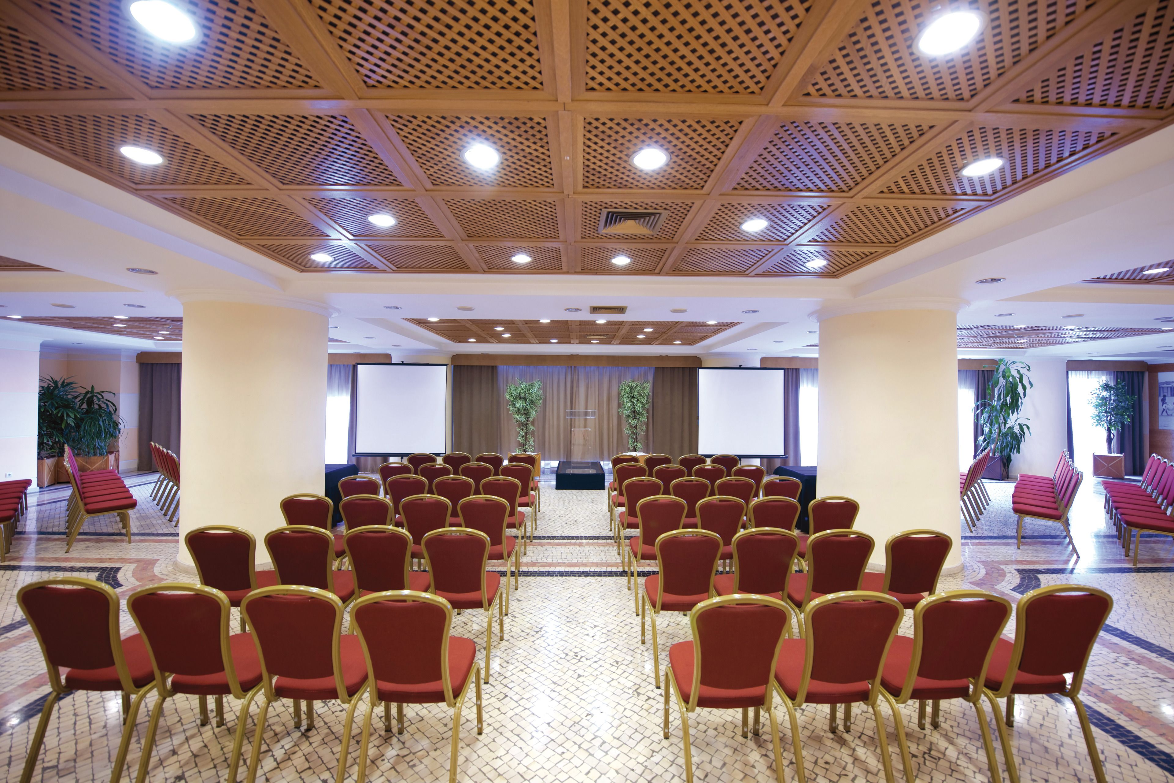 Une salle de conférence avec des rangées de chaises rouges, un pupitre et un grand écran à l’avant.