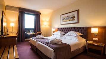 A hotel room with a large bed, a desk, a chair, a bedside table, and a window with curtains.
