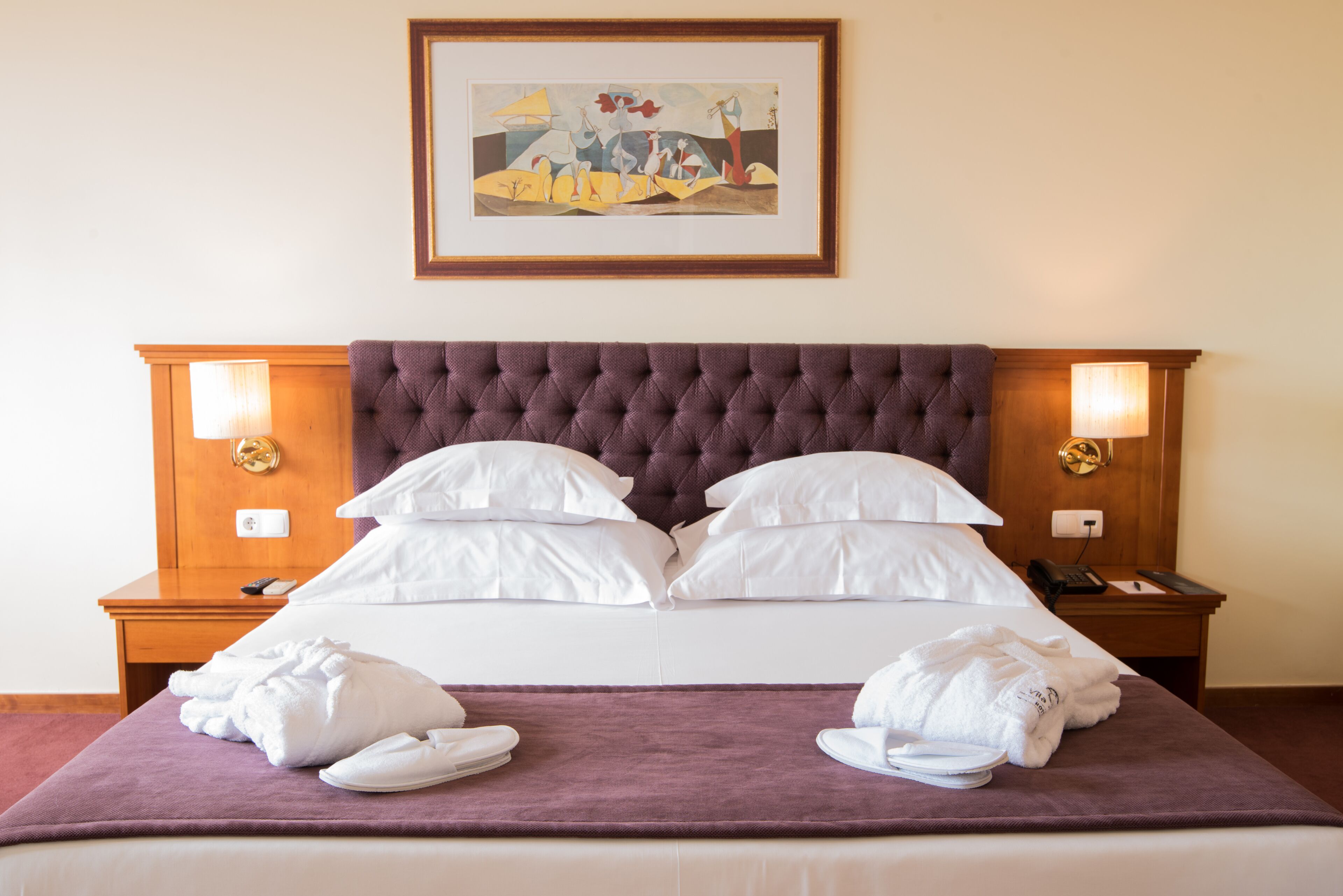 A neatly made bed with a tufted headboard, white pillows, and a purple blanket. There are two bedside tables with lamps and a framed artwork above the bed.