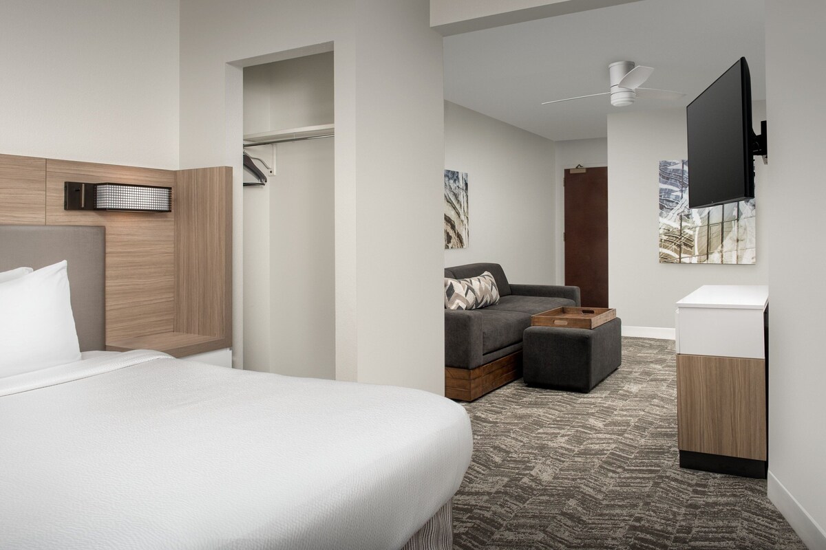 Een hotelkamer met een bed, een zithoek met een bank en voetenbankje, een aan de muur bevestigde televisie en een plafondventilator.