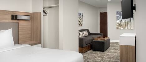 Una habitación de hotel con una cama, una zona de estar con sofá y reposapiés, un televisor montado en la pared y un ventilador de techo.