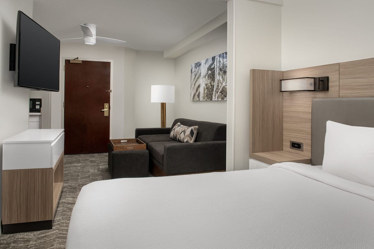 A hotel room with a bed, a sofa, a desk, a flat-screen TV, a ceiling fan, a wall-mounted lamp, and a painting.