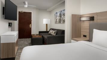 A hotel room with a bed, a sofa, a desk, a flat-screen TV, a ceiling fan, a wall-mounted lamp, and a painting.