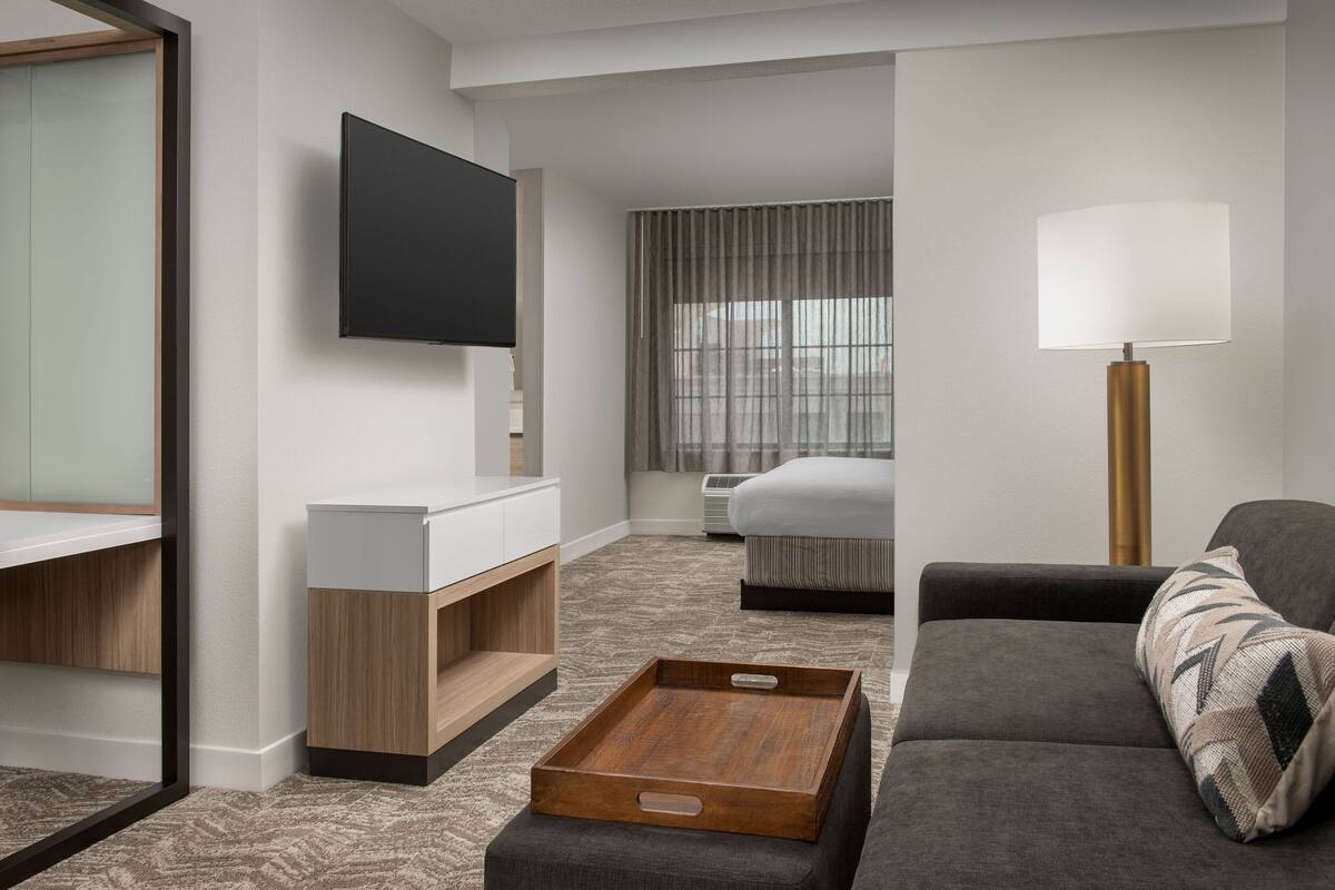 A modern hotel room with a flat-screen TV, a bedside table, a sofa, a lamp, and a window with curtains.