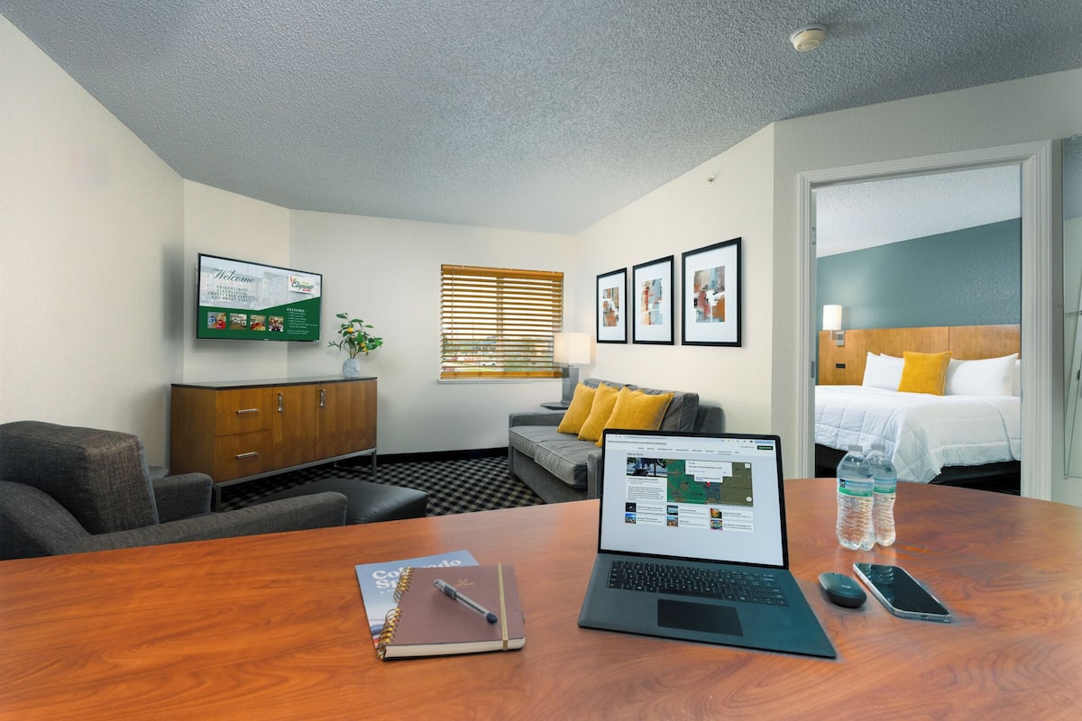 A hotel room with a wooden desk, a laptop, a notebook, a smartphone, and a bed with a yellow pillow.