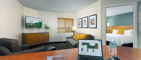 Une chambre d’hôtel avec un bureau en bois, un ordinateur portable, un carnet, un téléphone intelligent et un lit avec un oreiller jaune.