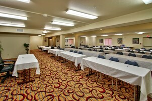 Un salón de conferencias con filas de mesas y sillas, suelo alfombrado e iluminación en el techo.