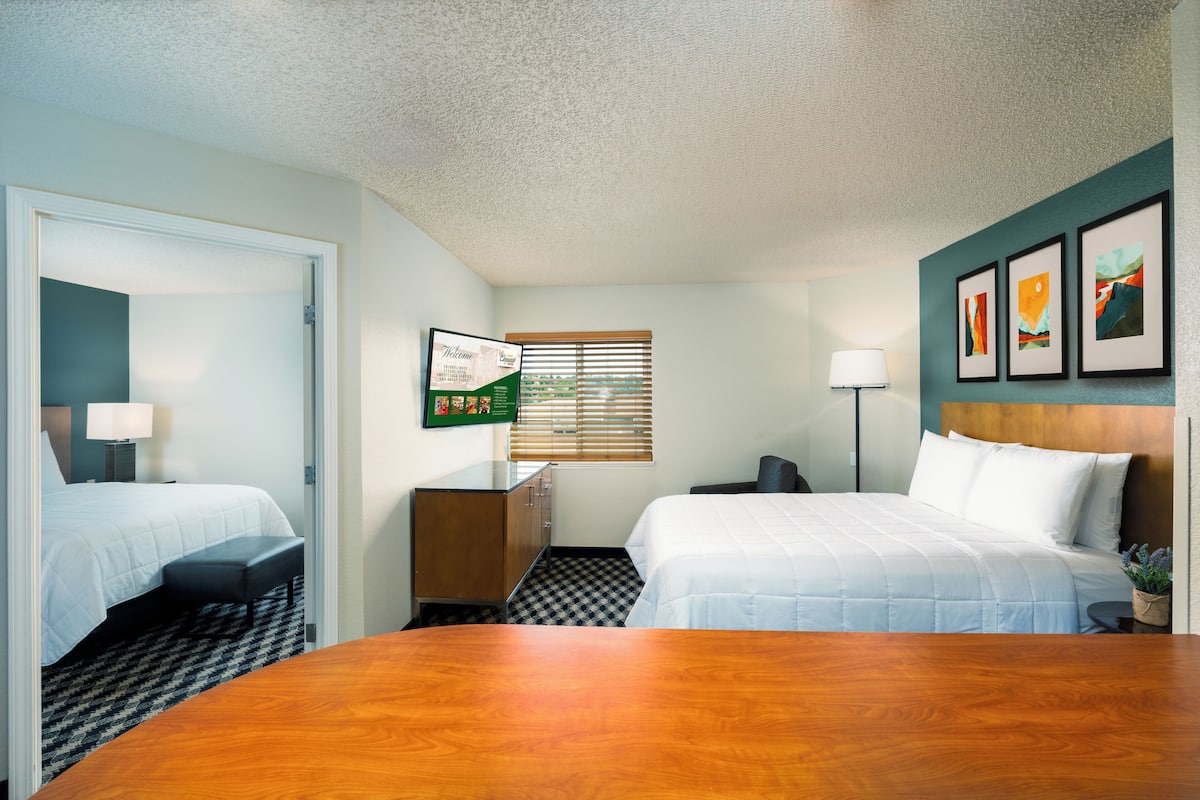 A hotel room with two beds, a desk, a chair, a lamp, a window with blinds, and artwork on the wall.