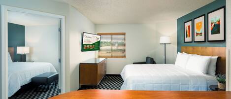 A hotel room with two beds, a desk, a chair, a lamp, a window with blinds, and artwork on the wall.
