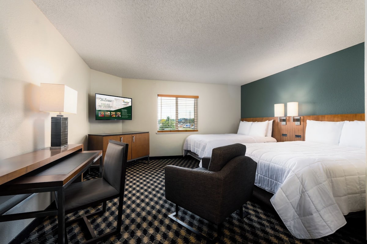 A hotel room with a desk, two beds, a chair, a television, and a window with blinds.