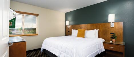 A hotel room with a bed, a nightstand, a lamp, a window with blinds, and a view of the outside.