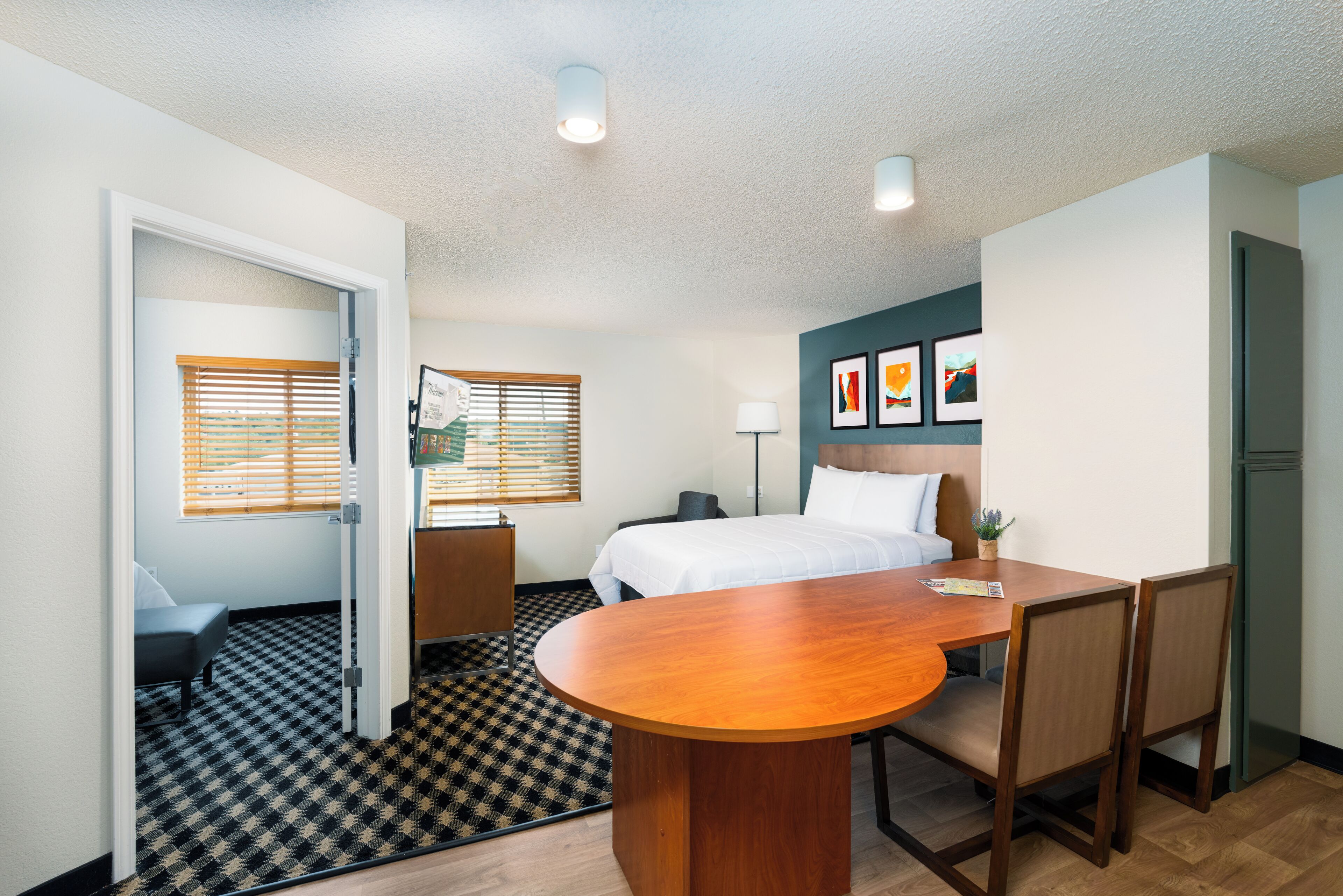 A hotel room with a bed, bedside table, desk, and chairs. There is a window with blinds, a lamp, and framed pictures on the wall.