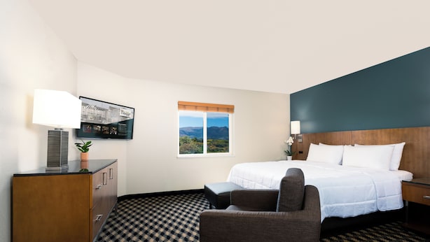 A hotel room with a bed, a desk, a chair, a TV on the wall, and a window with a mountain view. - MCM Elegante Suites (Colorado Springs)