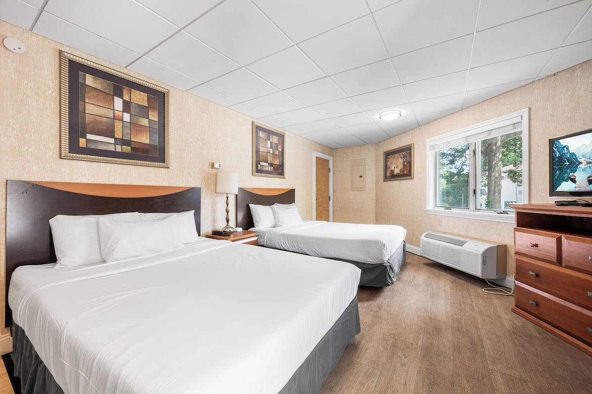Ein Hotelzimmer mit zwei Betten, einem Schminktisch, einem Fernseher und einem Fenster mit Blick auf Bäume.