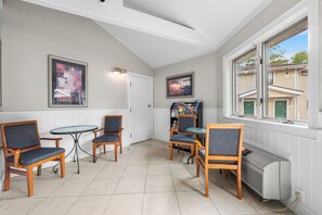 A corner room with a white wall, two framed pictures, a small round table with four chairs, a radiator, and a window with a view of houses. - Cedar Park Whirlpool Suites (North Stonington)