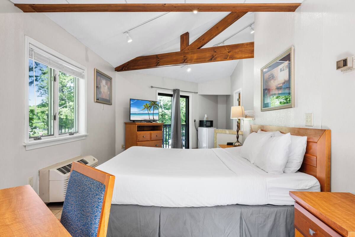 Ein Schlafzimmer mit einem großen Bett, Holzbalken, einem Schreibtisch, einem Fernseher und Blick auf Grünfläche.