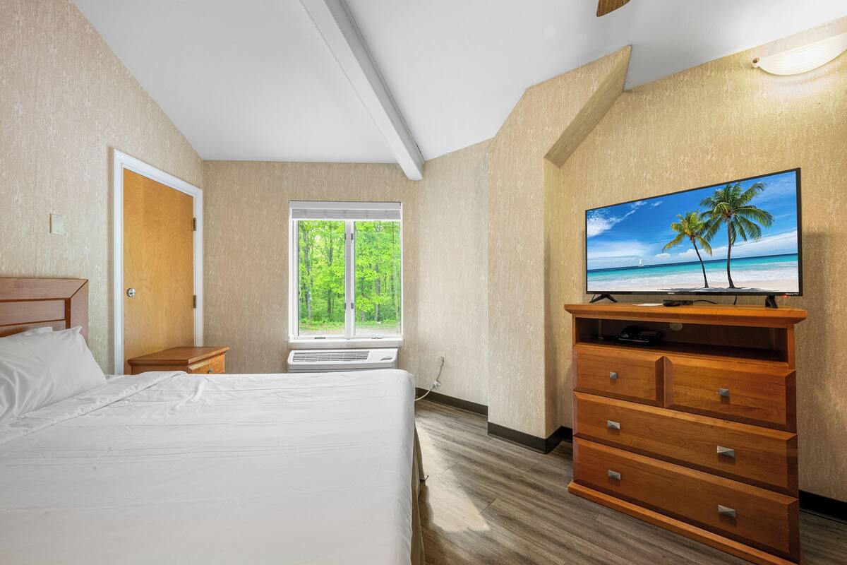 Ein Hotelzimmer mit Bett, einem Holzschrank, einem Fernseher und einem Fenster mit Blick auf Bäume und einen Strand.