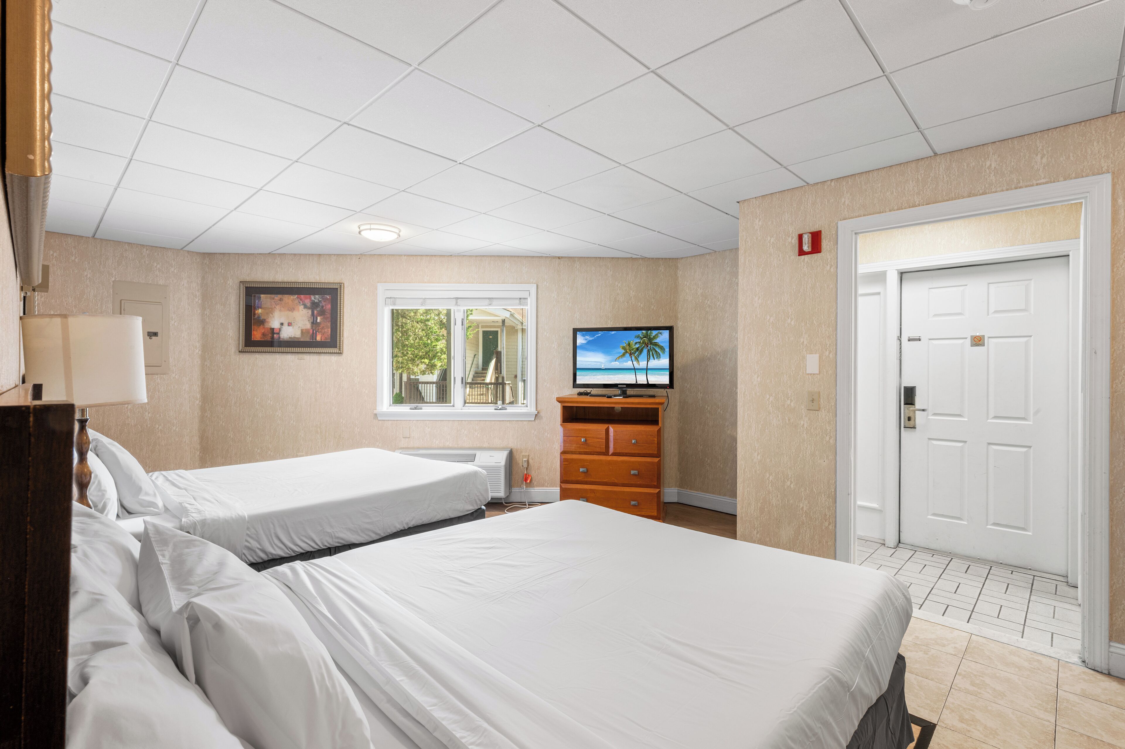 A hotel room with two beds, a television, a dresser, a door, and a window with a view of trees.